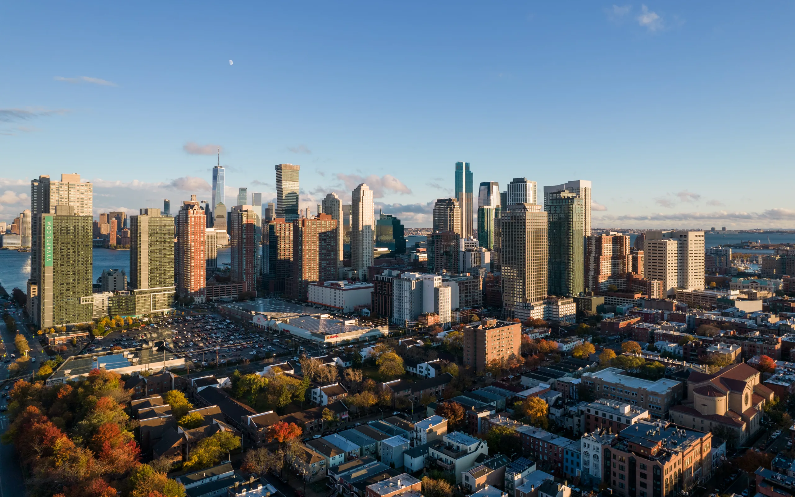 Sunset aerial over the neighborhood's autumn rooftops toward the Downtown towers and the Hudson